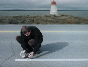 photograph of a man painting a white stripe on a road. the man is in front of a light house on a pier