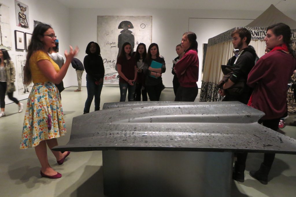 A group of people gathered around an art exhibition listening to an instructor