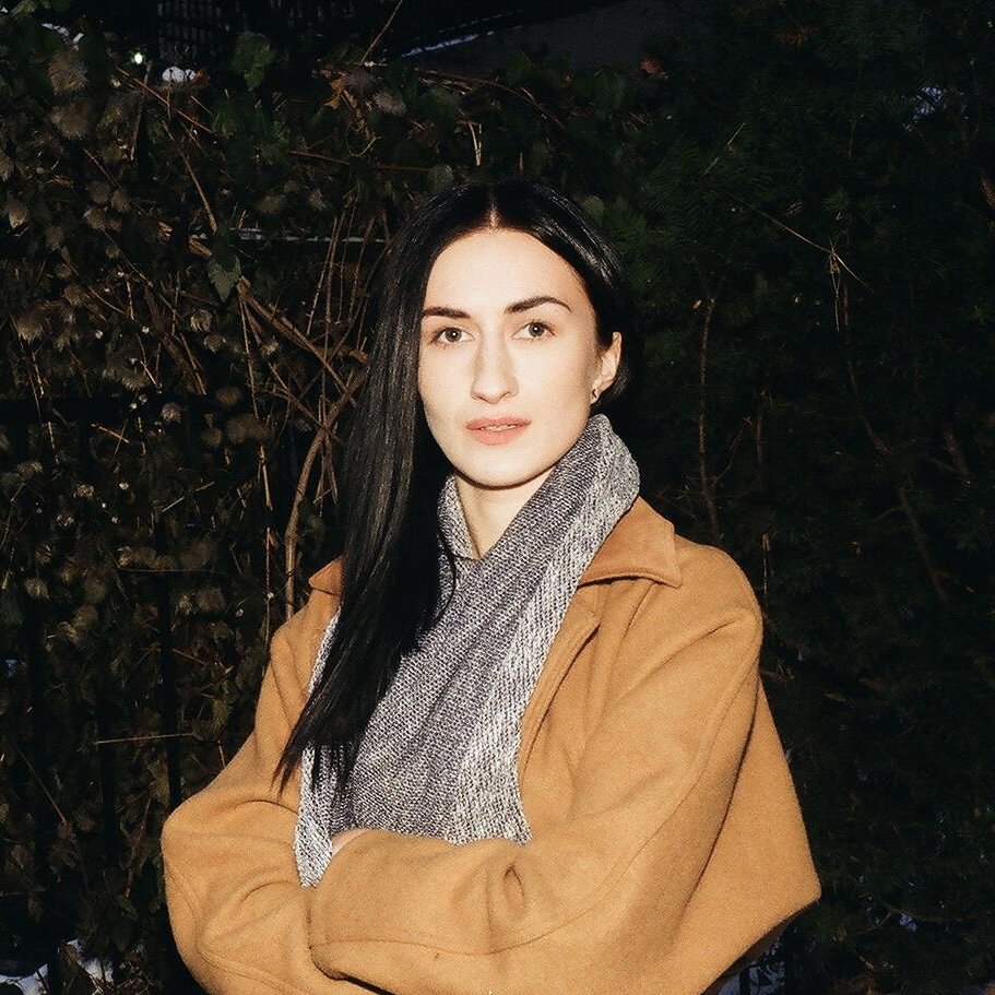 Portrait of Olga Kolotylo looking straight at the camera. Olga is wearing a beige jacket, grey scarf, and long black hair. Their arms are crossed and they stand in front of a shrub.