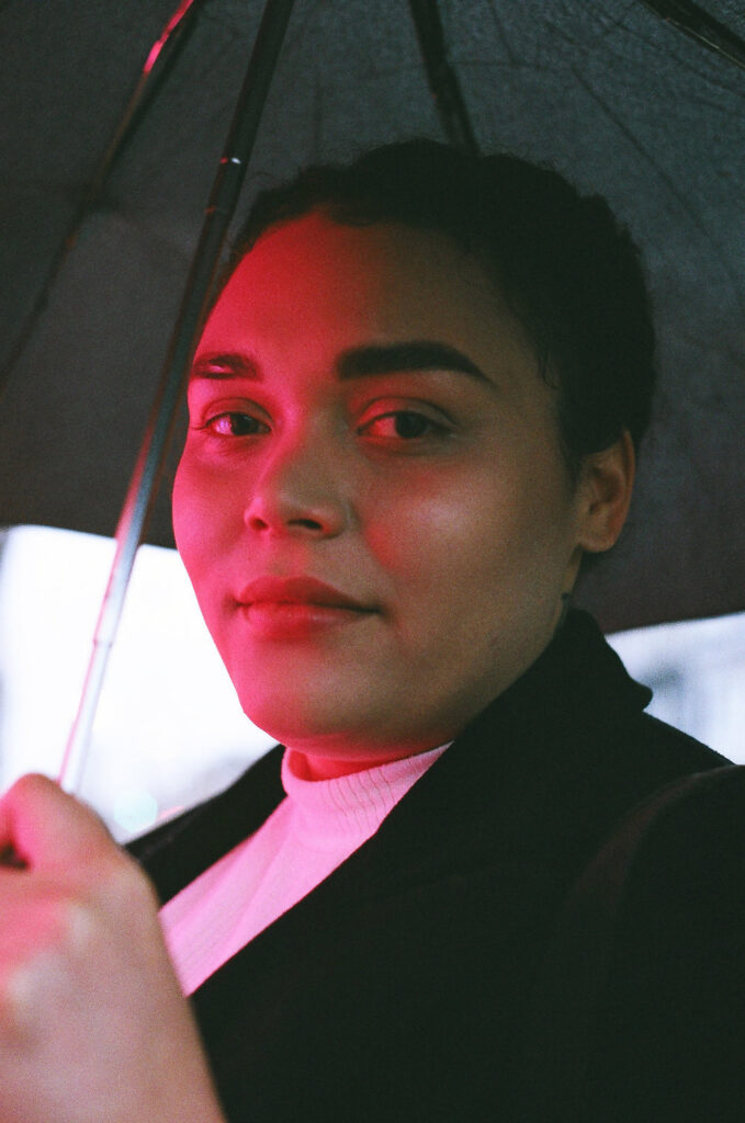 A portrait of Sarah-Tai Black looking straight at the camera. They are wearing a white shirt and black jacket and have short black hair. They hold a black umbrella behind their head and a soft pink glow lights their face.