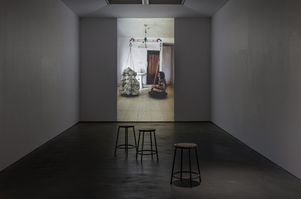 Installation view of Scapegoat - a video still of the artist sitting on a metal pan of a scale, facing a collection of bones of the same weight wrapped in cloth. the artist sits in a generic room with a sink and door. In the foreground are stools scattered about the museum space.