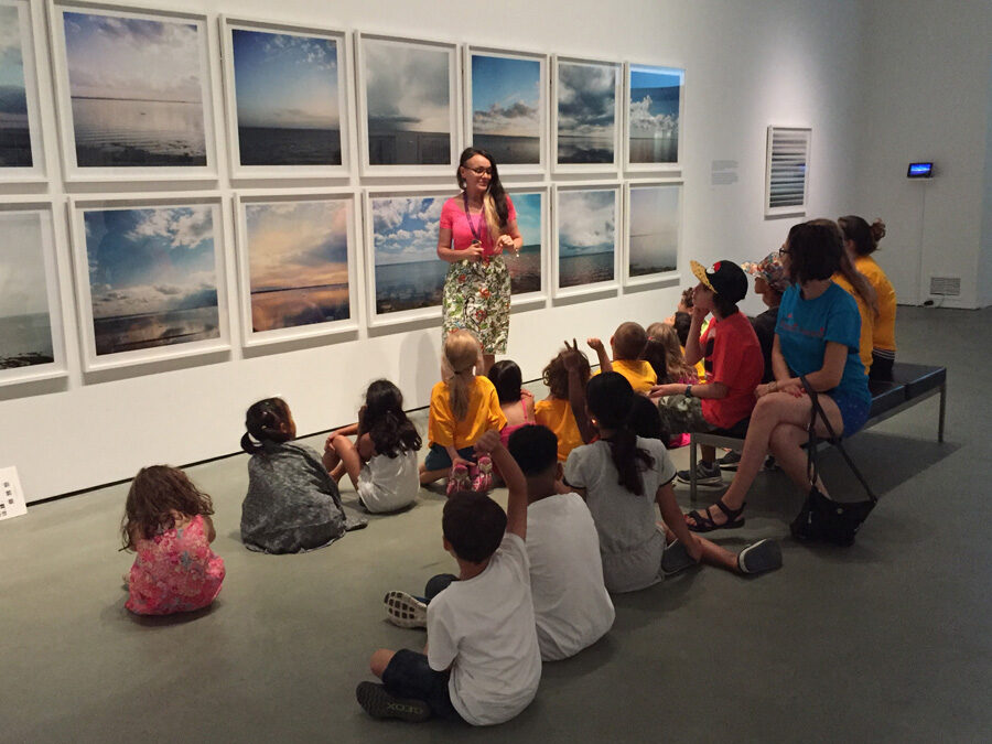 Person standing in front of a series of photographs speaking to a group of kids sitting on the floor