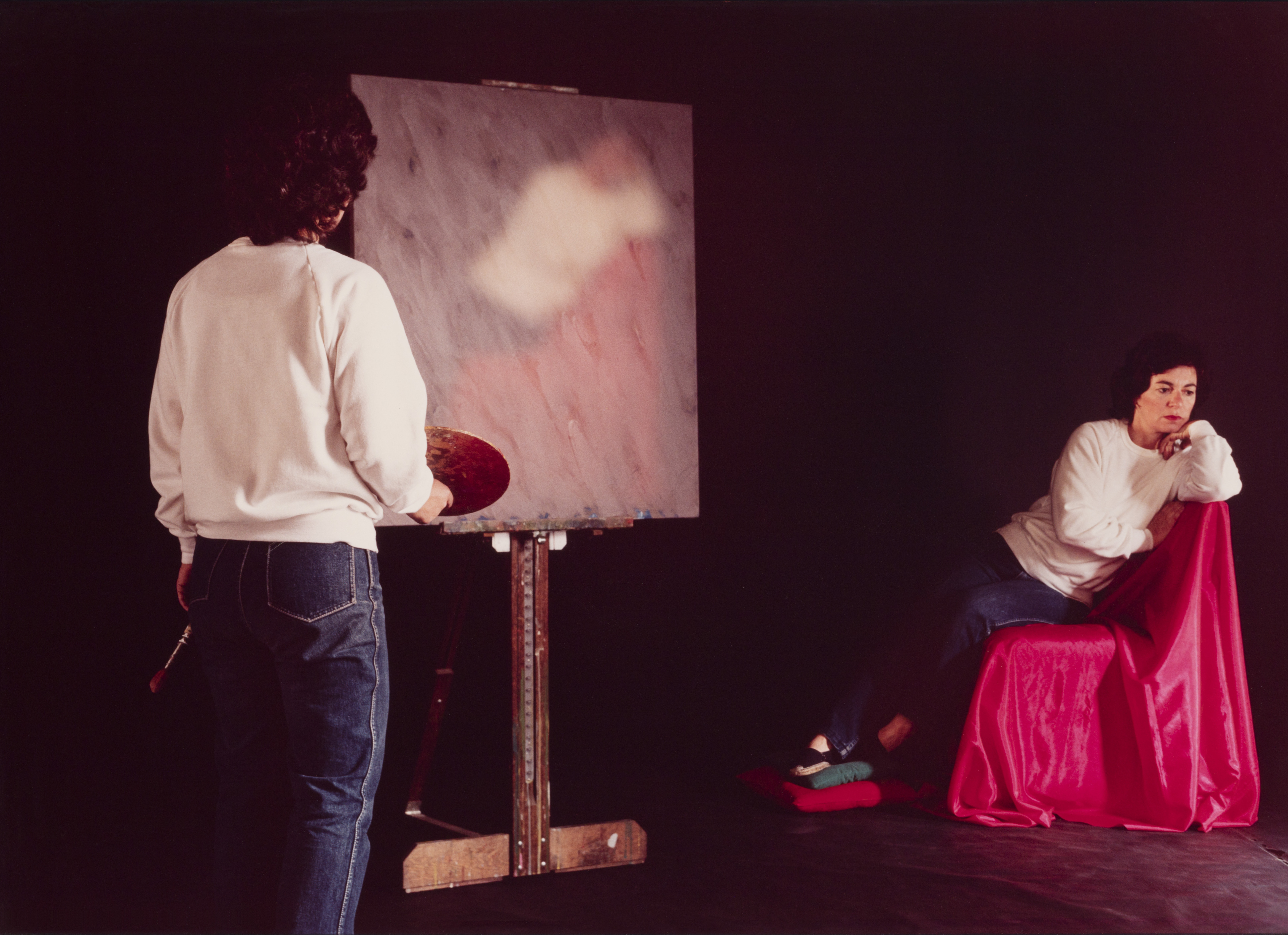 A person in a white sweater and jeans stands in front of a canvas with a paint palette. Beside them on the right is their subject in the same outfit posed on a red draped chair.