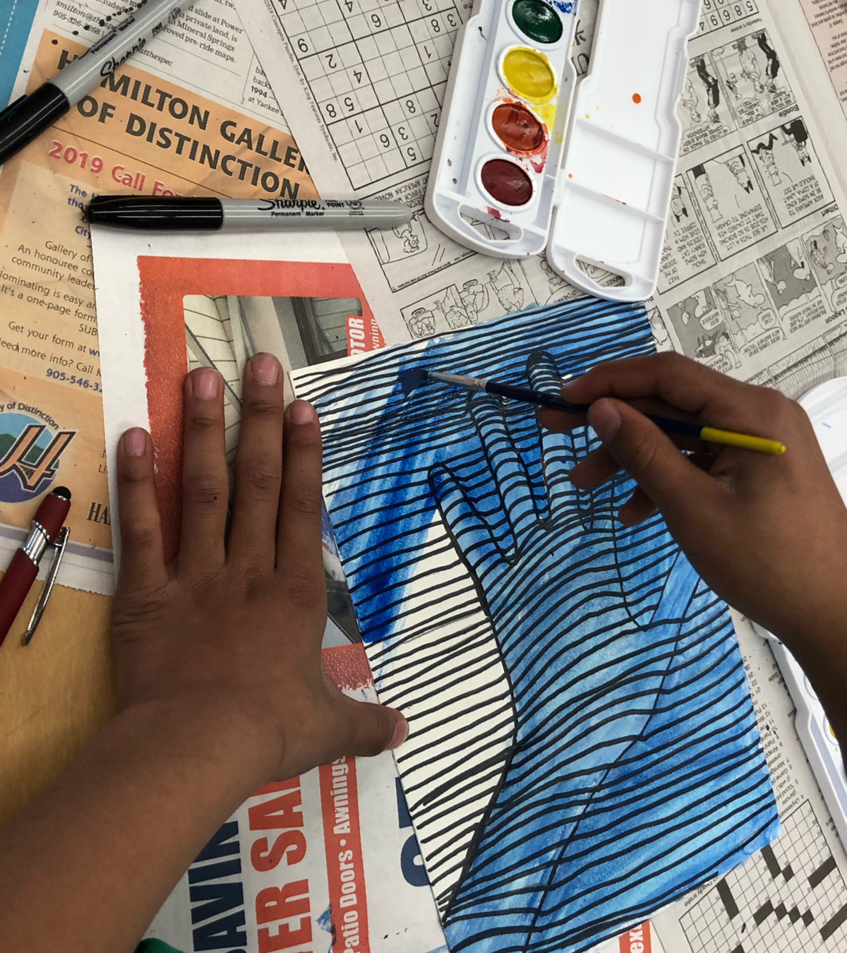 Student hands painting a striped artwork of a hand.