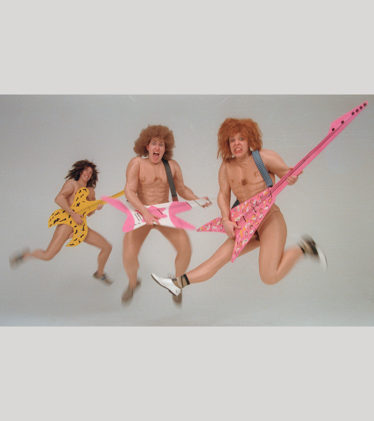 A photo of The Clichettes dressed as muscular men with wild hair, they are jumping mid-air holding colourful guitars.