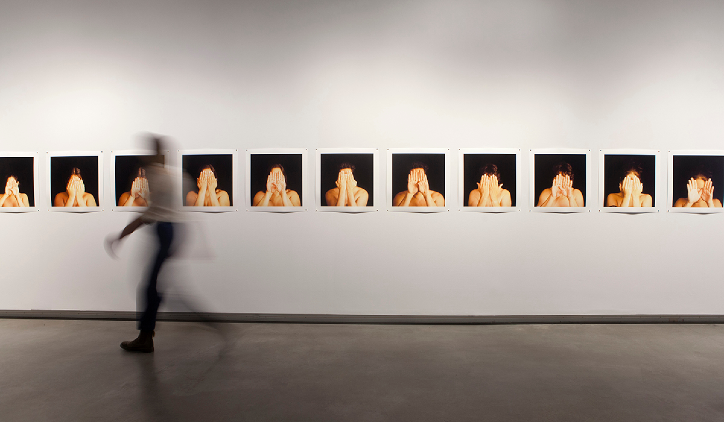 A blurred person walks in front of a wall featuring the photographic series Grace (2006). The artworks on display are photos of figures with hands covering their faces. Installation view of I'm Not Your Kind of Princess at Plug In ICA.